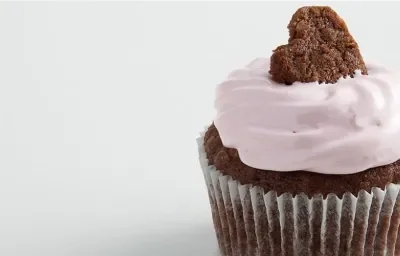 Fotografia em tons de branco, marrom e rosa vista de frente. Contém um bolinho de chocolate com creme de cereja e para decorar um mini bolinho em formato de coração por cima. Fotografia em tons de branco, marrom e rosa vista de frente. Contém um bolinho de chocolate com creme de cereja e para decorar um mini bolinho em formato de coração por cima.