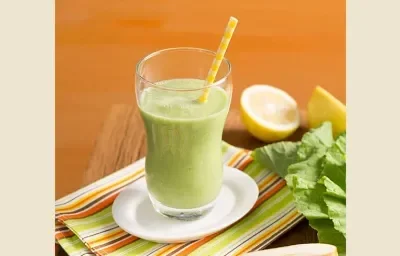 Fotografia em tons de verde e laranja em uma mesa de madeira com um pano listrado colorido e um copo de vidro com o suco verde de pera, couve e gengibre em cima dele. Ao lado, uma laranja cortada ao meio e folhas de couve.