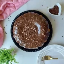 Foto em tons de marrom da receita do melhor bolo de cenoura servido sobre uma base preta com bastante cobertura de brigadeiro por cima com um paninho cor de rosa ao lado