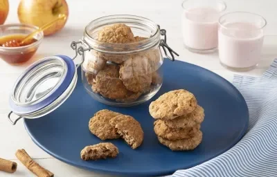 Foto da receita de biscoitos de maçã com iogurte nesfit servida em diversas porções, algumas dentro de um pote de vidro hermético e outras sobre um prato azul escuro com dois copos de iogurte nesfit ao fundo além de um vidro de mel