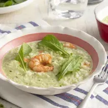 Fotografia em tons de verde e azul de uma bancada, ao centro um prato fundo com o risoto esverdeado de rúcula, sobre ele um camarão e 3 folhas de rúcula. Ao lado uma travessa vermelha e um garfo.