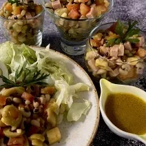 Foto da receita de Salada de Feijão Fradinho. Observa-se um prato grande com a salada e, ao lado, três potes individuais de vidro com a receita. Ao lado de tudo, um potinho com o molho de mostarda.