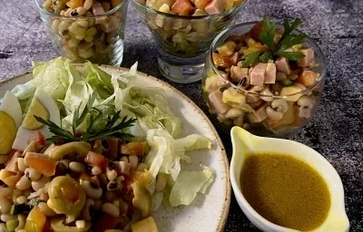 Foto da receita de Salada de Feijão Fradinho. Observa-se um prato grande com a salada e, ao lado, três potes individuais de vidro com a receita. Ao lado de tudo, um potinho com o molho de mostarda.