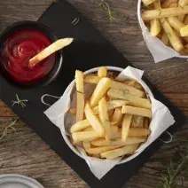 Fotografia em tons de marrom em uma bancada de madeira escura, uma tábua de ferro preta com um pote com batatas fritas dentro e um potinho ao lado com ketchup. Ao lado, um paninho e mais batata frita.
