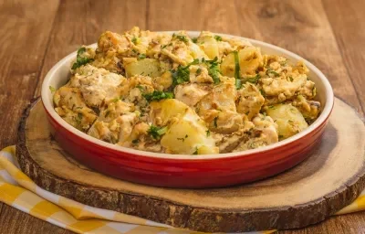 Foto de uma bancada de madeira. Ao centro há uma tábua de madeira sobre um pano quadriculado amarelo e branco e, sobre ela, um prato vermelho com a receita de frango e batatas gratinadas Foto de uma bancada de madeira. Ao centro há uma tábua de madeira sobre um pano quadriculado amarelo e branco e, sobre ela, um prato vermelho com a receita de frango e batatas gratinadas