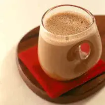 Fotografia em tons de vermelho em uma bancada de madeira, uma tábua de madeira escura, um paninho vermelho, uma xícara de vidro com a bebida quente feita com chocolate em pó.