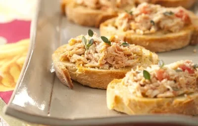 Fotografia em tons de bege em uma bancada de madeira clara com uma toalha rosa e amarela e um recipiente retangular bege com as bruschettas com o frango desfiado em cima dele.