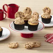 Foto de uma bancada clara com canecas e um bule vermelhos, um tecido listrado vermelho e branco. Há dois pratos brancos com cupcakes em cima e, ao centro, um prato com três cupcakes decorados com cookies.