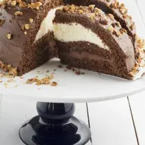 Fotografia em tons de marrom em uma bancada de madeira clara, um suporte de bolo branco com pé preto e uma doce de chocolate com recheio de creme branco e de chocolate.