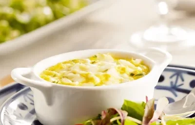 Fotografia em tons de verde e azul em uma bancada de madeira branca, um prato branco com detalhes azuis ao centro e salada verde em cima dele, com um potinho branco e a brandade de bacalhau dentro dele.