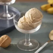 Foto da receita de Sorvete Crocante de Farinha Láctea Nestlé. Observa-se uma taça de sobremesa de vidro transparente com duas bolas do sorvete. A foto é decorada com Farinha Láctea, nozes com casca e bananas