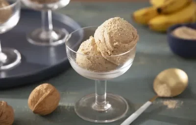 Foto da receita de Sorvete Crocante de Farinha Láctea Nestlé. Observa-se uma taça de sobremesa de vidro transparente com duas bolas do sorvete. A foto é decorada com Farinha Láctea, nozes com casca e bananas