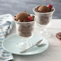 Fotografia em tons de azul de uma bancada branca, ao centro um pratinho azul com uma taça com duas bolas de sorvete de chocolate e uma cereja. Ao fundo outra taça com sorvete de chocolate e um potinho com raspas de chocolate.