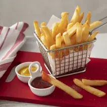 Fotografia em tons de vermelho de uma bancada branca, ao centro uma tábua vermelha com uma cestinha com batatas fritas dentro. Na frente um potinho com ketchup e um com mostarda.