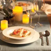 Fotografia em tons de amarelo em uma mesa de madeira e uma toalha de mesa cinza. Um sousplat branco e um prato fundo branco com a sofioli de shimeji ao molho de tomates. Velas amarelas, taças de vidro, arranjo pequeno de flor e talheres compondo a mesa.