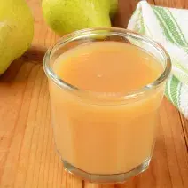 Fotografia em tons de marrom, laranja, branco e verde de uma bancada de madeira marrom vista de cima, contém um copo transparente com suco de pera e maçã. Ao fundo duas peras e ao lado um pano branco e verde