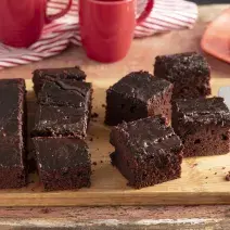 Fotografia em tons de marrom e vermelho de uma bancada de madeira, ao centro uma tábua com fatias de bolo de chocolate. Ao fundo 2 xícaras e um prato vermelho com uma fatia de bolo.