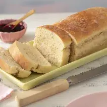 Receita de Pão de Leite Moça. Observa-se um pão cortado com três fatias sob uma forma retangular verde clara. Ao lado, uma faca de pão e do outro, um potinho com geleia.