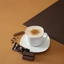 Foto da receita de cappuccino chai cremoso servida em uma xícara branca com um kit kat dark ao lado em fundo marrom claro e escuro
