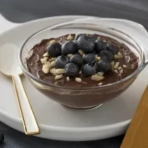 Foto em tons escuros da receita de mousse de chocolate com mirtilo servida em uma taça de vidro sobre uma frigideira de porcelana branca com uma colher branca ao lado