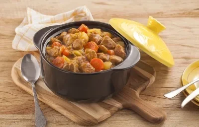 Fotografia em tons de amarelo em uma bancada de madeira clara, um pano amarelo com listras, uma tábua de madeira, uma panela preta de ferro com o picadinho de carne suína com legumes dentro e uma tampa amarela ao lado. Fotografia em tons de amarelo em uma bancada de madeira clara, um pano amarelo com listras, uma tábua de madeira, uma panela preta de ferro com o picadinho de carne suína com legumes dentro e uma tampa amarela ao lado.