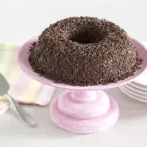 Foto de um brigadeirão de chocolate em cima de um prato de bolo rosa e com pratos brancos atrás