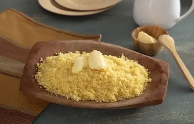 Foto da receita de Cuscuz de Milho. Observa-se uma tigela de madeira com o cuscuz soltinho e com manteiga em cima derretendo. Foto da receita de Cuscuz de Milho. Observa-se uma tigela de madeira com o cuscuz soltinho e com manteiga em cima derretendo.