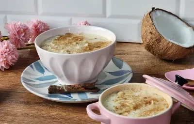 Foto da receita de cocada de panela de pressão, servida em uma tigela branca, sobre um prato decorado com pau de canela, numa bancada de madeira com um pedaço de coco seco aberto e algumas flores rosas de mentira ao fundo Foto da receita de cocada de panela de pressão, servida em uma tigela branca, sobre um prato decorado com pau de canela, numa bancada de madeira com um pedaço de coco seco aberto e algumas flores rosas de mentira ao fundo