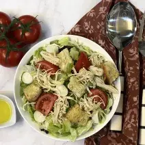 Foto vista de cima da receita de Salada de Frango servida em um recipiente grande, com um recipiente menor com Molho de Mostarda. Tudo está sobre uma bancada decorada com um tecido vermelho, um par de talheres e alguns tomates