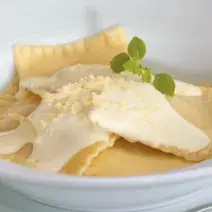 Fotografia em tons de amarelo em um fundo branco com um prato redondo grande, largo e branco com ravioli de abóbora com gorgonzola e molho branco em cima.
