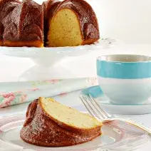 Fotografia em tons de azul em uma mesa branca com uma toalha branca, um prato de sobremesa com uma fatia de bolo de maracujá. Ao fundo, um suporte para bolo com o bolo inteiro e ao lado uma xícara com listra azul e chá dentro dela.