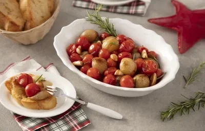 Foto da receita de Confit de Tomates e Cebolas. Observa-se um refratário branco de porcelana com os tomates e as cebolas dentro, acompanhados de alho e alecrim. Ao lado esquerdo, um pratinho com uma torrada acompanhada do confit.