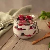 Foto da receita de overnight oats de frutas vermelhas servida em um pote de vidro hermético sobre uma mesa de madeira. Ao lado há um pano verde, uma colher de madeira e um pote de vidro com mel