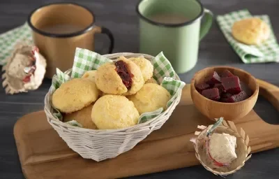 Foto em tons de amarelo e marrom da receita de pão de queijo romeu e julieta servida em diversas porções em uma cestinha sobre uma tábua de madeira com decoração de festa junina e um bowl com goiabada. Ao fundo há duas canecas