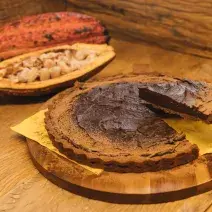 Fotografia em tons de marrom de uma bancada de madeira com uma tábua de madeira, sobre ela um paninho amarelo com uma torta de chocolate e uma espátula retirando uma fatia. Ao fundo um cacau.
