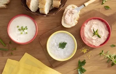 Foto da receita de trio de pates e ricota com maggi meu segredo servida em três porções com os diferentes patês em potinhos vermelhos e amarelos. Tudo isso sobre uma mesa de madeira decorada com pães e ervas