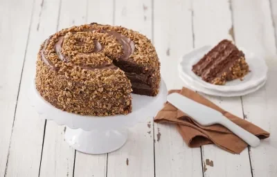 Fotografia em tons de marrom em uma mesa de madeira com um suporte para bolo branco ao centro e o bolo crocante de chocolate em cima. Ao lado, um guardanapo de pano marrom com uma espátula de bolo branco apoiada e um pratinho branco com uma fatia do bolo.