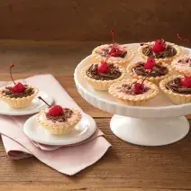 Fotografia em tons de vermelho em uma bancada de madeira, um paninho rosa, um suporte para bolo branco com várias tortinhas de Moça com cerejas para enfeitar. Ao lado, dois pratinhos brancos pequenos cada um com uma tortinha em cima.