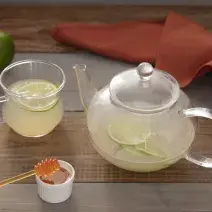 Fotografia em tons de verde em uma bancada de madeira escura, um bule de vidro com o chá de gengibre e limão dentro. Ao lado, uma xícara com o chá e uma fatia de limão e um potinho com mel. Gengibre e limões espalhados pela mesa.