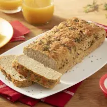Foto da receita de pão de forma sem glúten e sem lactose, fatiado sobre um prato retangular branco, em uma mesa decorada com um prato amarelo, fatias de pão, copos de suco de laranja