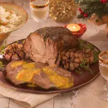 Fotografia em tons de amarelo em uma bancada de madeira, um pano bege, um prato redondo marrom com a copa lombo com o molho em cima dele. Ao fundo, um potinho com arroz e decoração e enfeites natalinos.