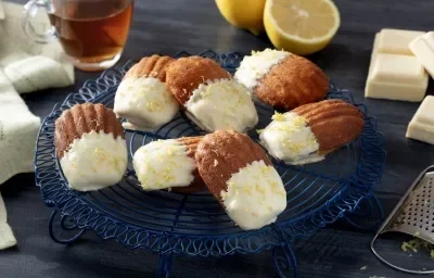 Fotografia em tons de azul em uma bancada de madeira de cor cinza. Ao centro, um prato azul marinho contendo as madeleines. Ao fundo, há uma xícara com mel, um limão partido e barras de chocolate branco. Fotografia em tons de azul em uma bancada de madeira de cor cinza. Ao centro, um prato azul marinho contendo as madeleines. Ao fundo, há uma xícara com mel, um limão partido e barras de chocolate branco.