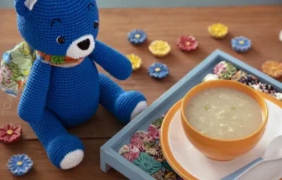 Fotografia em tons de branco em uma bancada de madeira de cor marrom. Ao centro, uma tábua azul clara contendo um prato amarelo em cima e um mini bowl com o creme. Ao lado, um ursinho azul e ao redor, há várias flores espalhadas.