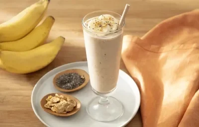 Foto da receita de Smoothie de Banana com Nesfit Cereal. Observa-se um copo alto com o smoothie dentro e finalizado com NESFIT Cereal, banana e chia. Ao lado esquerdo, potinhos com NESFIT cereal, chia e um cacho de bananas decoram a foto.