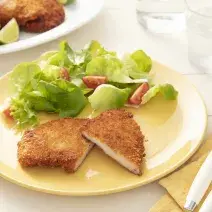 Fotografia em tons de amarelo de uma bancada branca, ao centro um prato amarelo com um filé de frango empanado cortado ao meio e salada de alface e tomate cereja. Ao lado um garfo e uma faca sobre um pano amarelo.