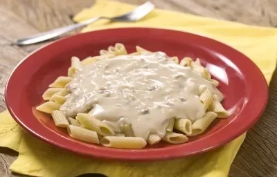Fotografia em tons de marrom, amarelo e vermelho de uma bancada de madeira com um paninho amarelo, sobre ele um prato vermelho com macarrão. Ao fundo um garfo.