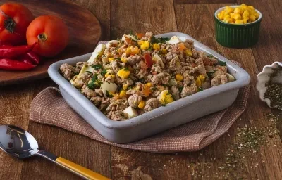 Imagem de uma bancada de madeira escura, sobre a qual há uma prato retangular em tom de cinza com a receita de refogado de carne suína, uma tábua de madeira com tomates em cima e ao fundo um pote pequeno com milho Imagem de uma bancada de madeira escura, sobre a qual há uma prato retangular em tom de cinza com a receita de refogado de carne suína, uma tábua de madeira com tomates em cima e ao fundo um pote pequeno com milho