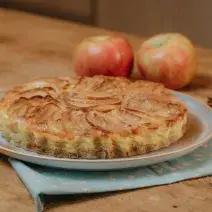 Fotografia em tons de marrom e azul de uma bancada de madeira com um paninho azul sobre ele um prato azul com uma torta de maçã. Ao fundo duas maçãs e paus de canela.