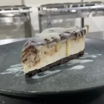 Foto da receita de CHEESECAKE MOÇA GANACHE. Observa-se uma fatia da cheesecake em um prato de cerâmica preto, com Leite MOÇA escorrendo pelo recheio.