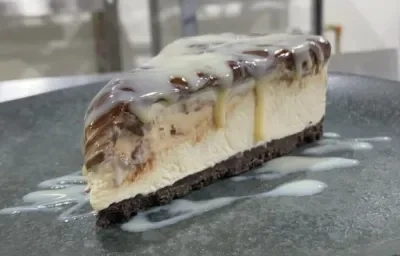 Foto da receita de CHEESECAKE MOÇA GANACHE. Observa-se uma fatia da cheesecake em um prato de cerâmica preto, com Leite MOÇA escorrendo pelo recheio.